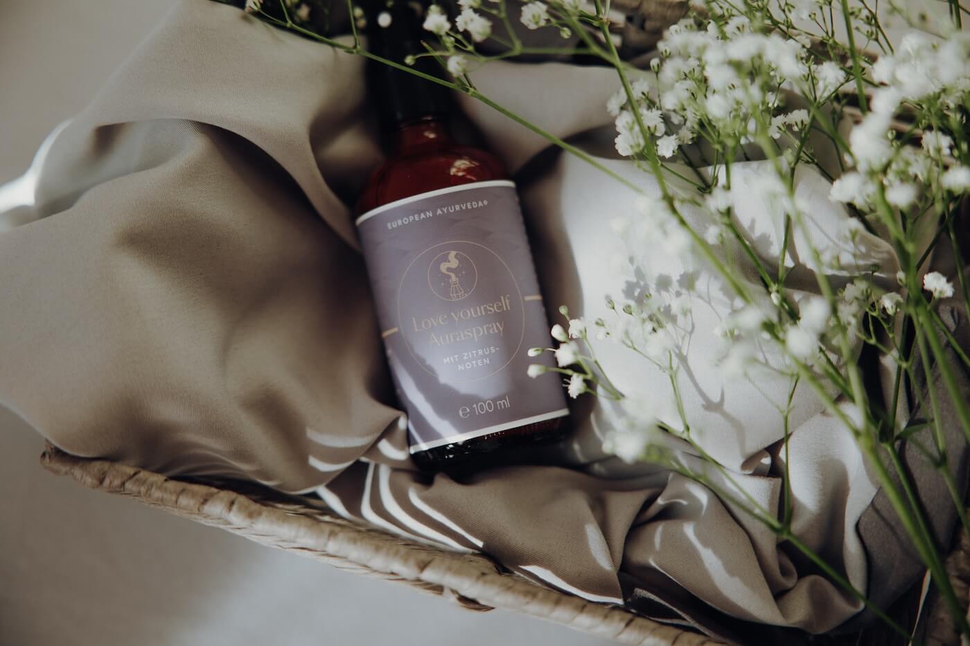 Raumduftspray liegt neben Blumen auf Stoff – Darstellung eines natürlichen Ayurveda Dufterlebnisses.