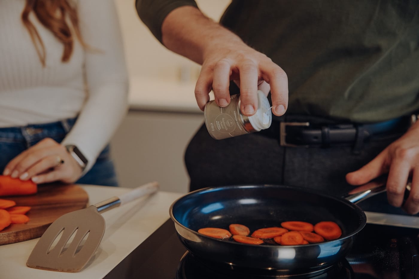 Hand würzt Gemüse in einer Pfanne – Darstellung von ayurvedischer Ernährung und gesunder Zubereitung von Lebensmitteln.