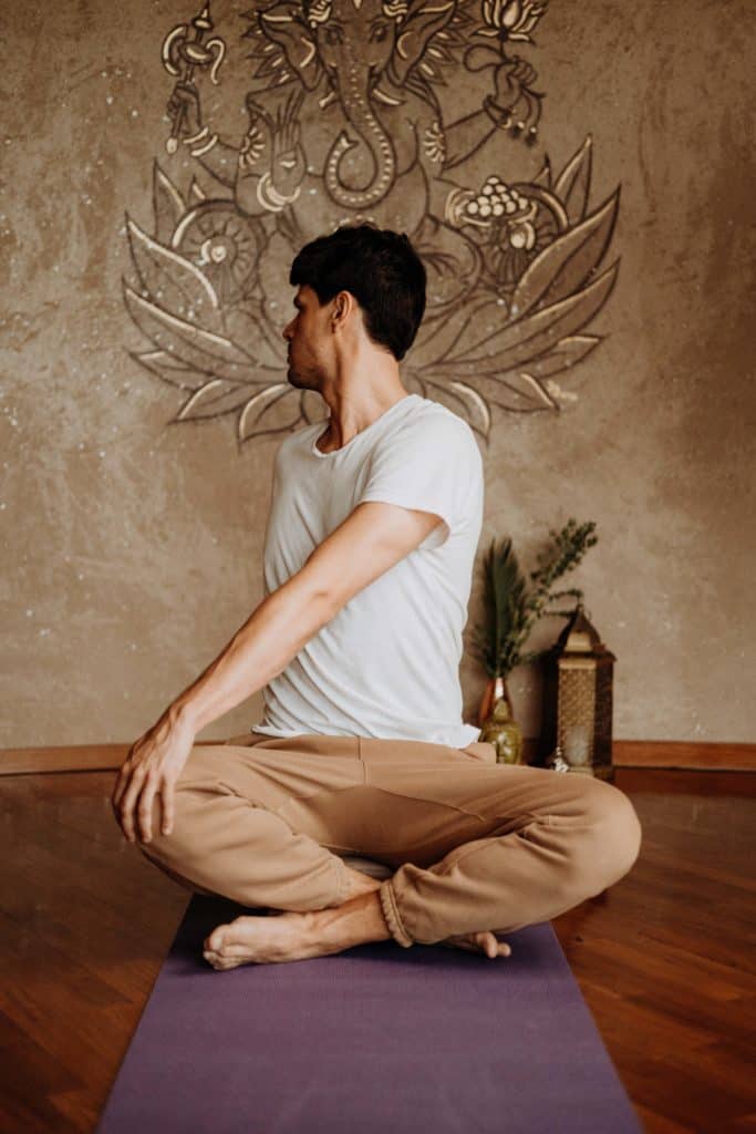 Ein Mann sitzt mit verschränkten Beinen auf einer Yoga-Matte und sein Oberkörper ist nach hinten gedreht